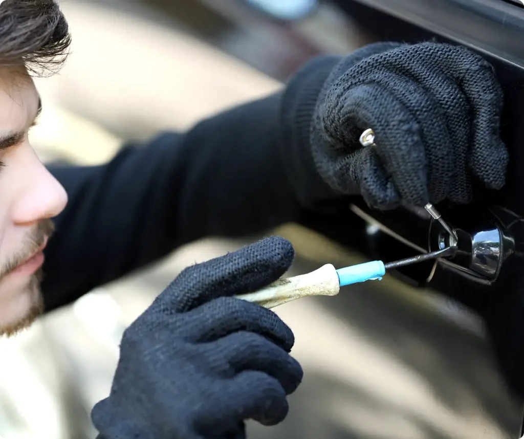 Osoba w czarnych rękawiczkach próbuje otworzyć drzwi samochodu za pomocą narzędzi. Scena sugeruje niezgodne z prawem działanie, wywołując napięcie i ostrożność.